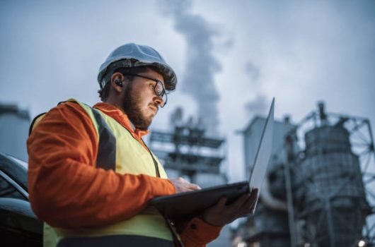 A shot of an young engineer wearing a helmet and using a laptop and hands free device during his night shirt in the oil rafinery. Engineering concept.
