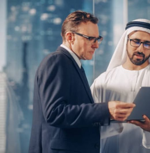 International Operations Manager Meeting Saudi Business Partner in Traditional Kandura. They're Standing in Modern Office, Using Laptop Computer. Successful Saudi, Emirati, Arab Businessman Concept.